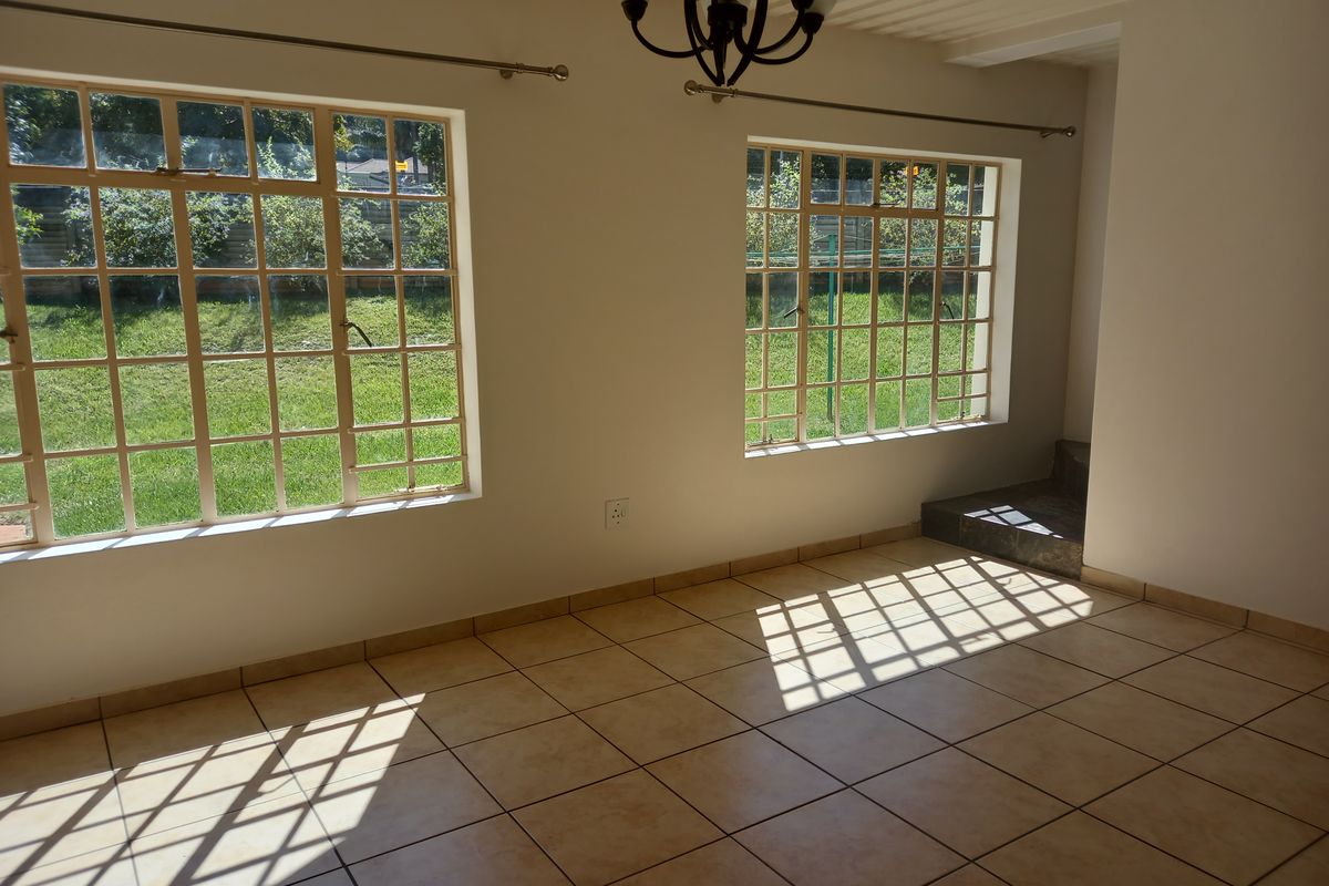 A bright, empty living area featuring neutral tiled flooring and large steel-framed windows overlooking the garden.