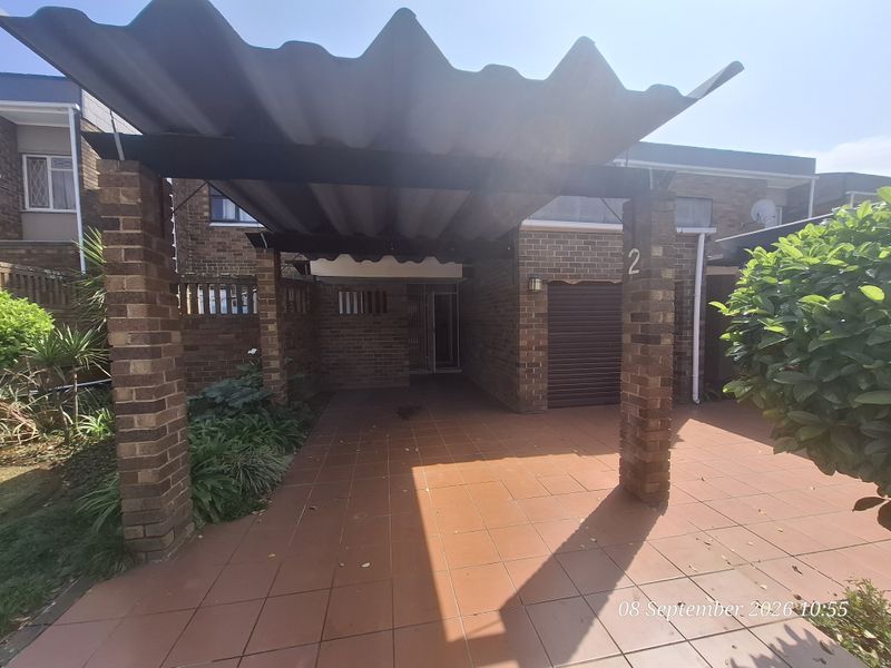 Front exterior view of a townhouse featuring a covered carport with brick pillars, tiled driveway, and a roller garage door.