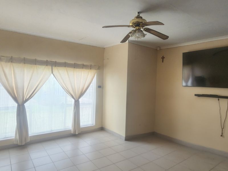 A spacious, empty living room featuring neutral tiled flooring, a ceiling fan, and a large window allowing for natural light.