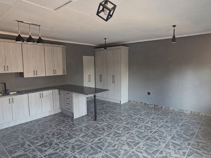 A modern, empty kitchen featuring light wood-grain cabinetry, dark laminate countertops, a functional island, and tiled flooring with contemporary pendant lighting.