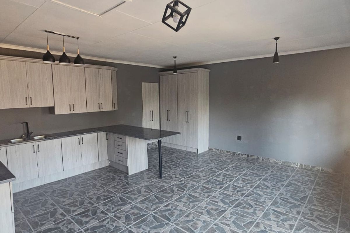 A modern, empty kitchen featuring light wood-grain cabinetry, dark laminate countertops, a functional island, and tiled flooring with contemporary pendant lighting.