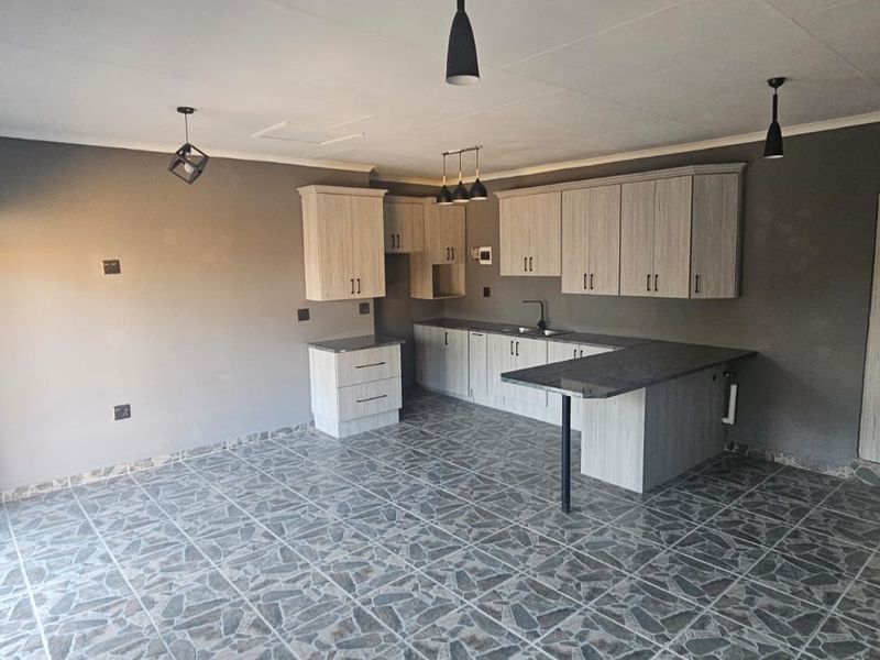 A modern, open-plan kitchen featuring light wood-grain cabinetry, dark countertops, tiled flooring, and contemporary pendant lighting in a clean, vacant space.