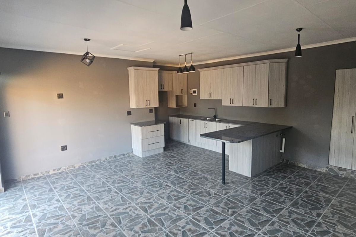A modern, open-plan kitchen featuring light wood-grain cabinetry, dark countertops, tiled flooring, and contemporary pendant lighting in a clean, vacant space.
