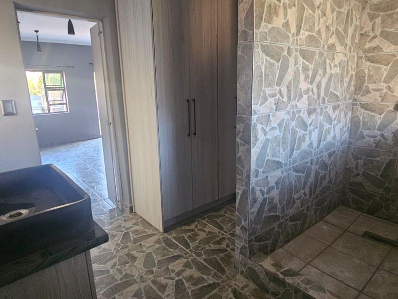 A modern bathroom featuring a walk-in shower with stone-look tiling, a vessel sink vanity, and built-in cupboards, with an adjacent view into another room.