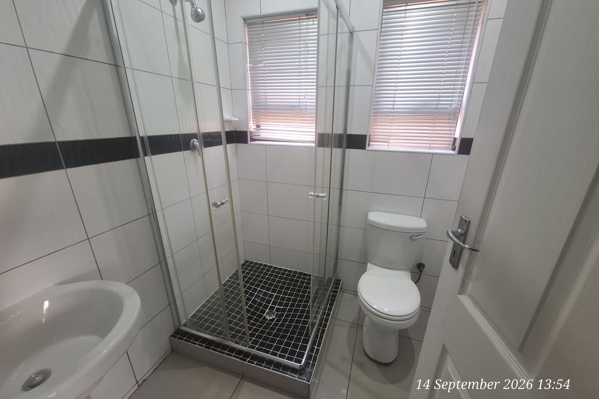 A clean, functional bathroom featuring a glass-enclosed shower, pedestal basin, and toilet with neutral wall and floor tiling.