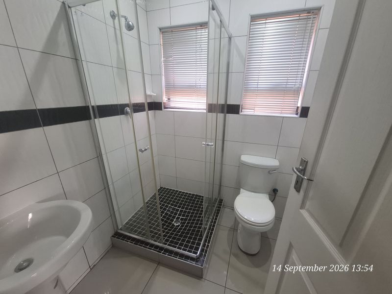 A clean, functional bathroom featuring a glass-enclosed shower, pedestal basin, and toilet with neutral wall and floor tiling.