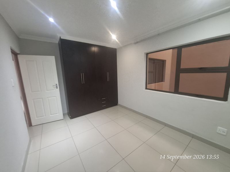 A clean, empty bedroom featuring neutral tiled flooring, built-in dark wood cupboards, and modern recessed downlighting.