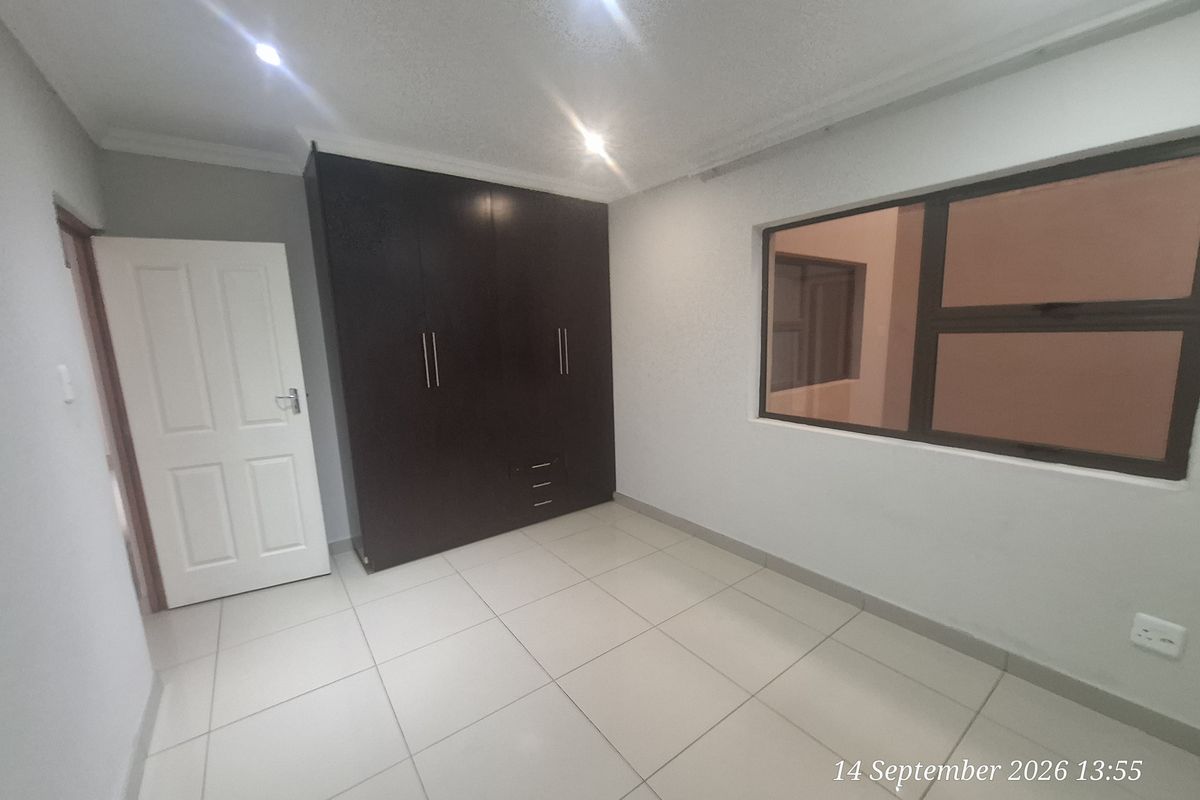 A clean, empty bedroom featuring neutral tiled flooring, built-in dark wood cupboards, and modern recessed downlighting.