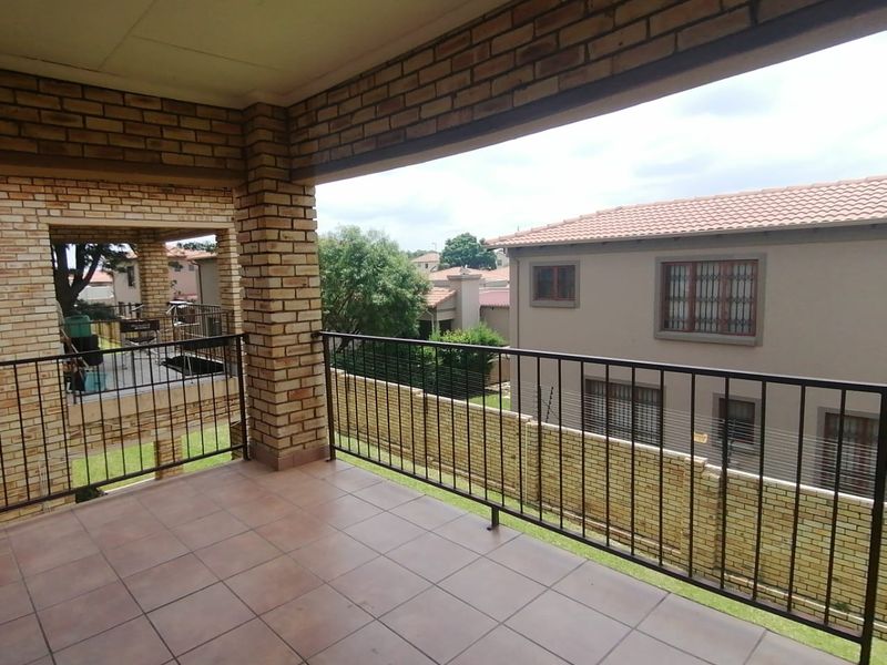 A spacious covered balcony featuring tiled flooring, exposed brick pillars, and a metal balustrade, offering an open view of the surrounding townhouse complex.
