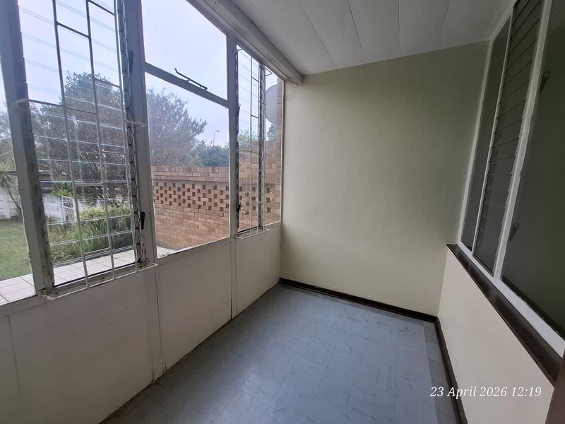 An enclosed patio or sunroom featuring large windows with security bars and tiled flooring, offering a bright space overlooking the garden area.
