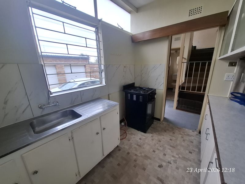 A dated kitchen featuring a stainless steel sink, gas stove, and original cabinetry, with direct access to an internal staircase.