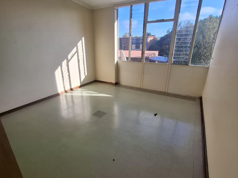 An empty, sunlit bedroom featuring large windows with louver sections, a central pendant light, and vinyl flooring, ready for renovation or staging.