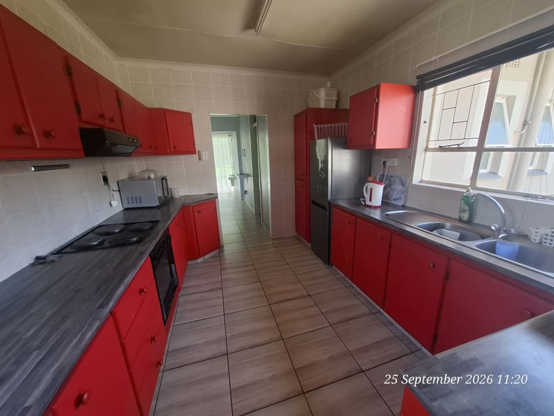 A functional kitchen featuring red cabinetry, dark countertops, a built-in electric hob, oven, and stainless steel sink with tiled flooring and backsplash.