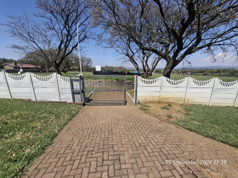 A paved walkway leading to a metal security gate and precast concrete boundary wall, set against a backdrop of mature trees and open landscape.