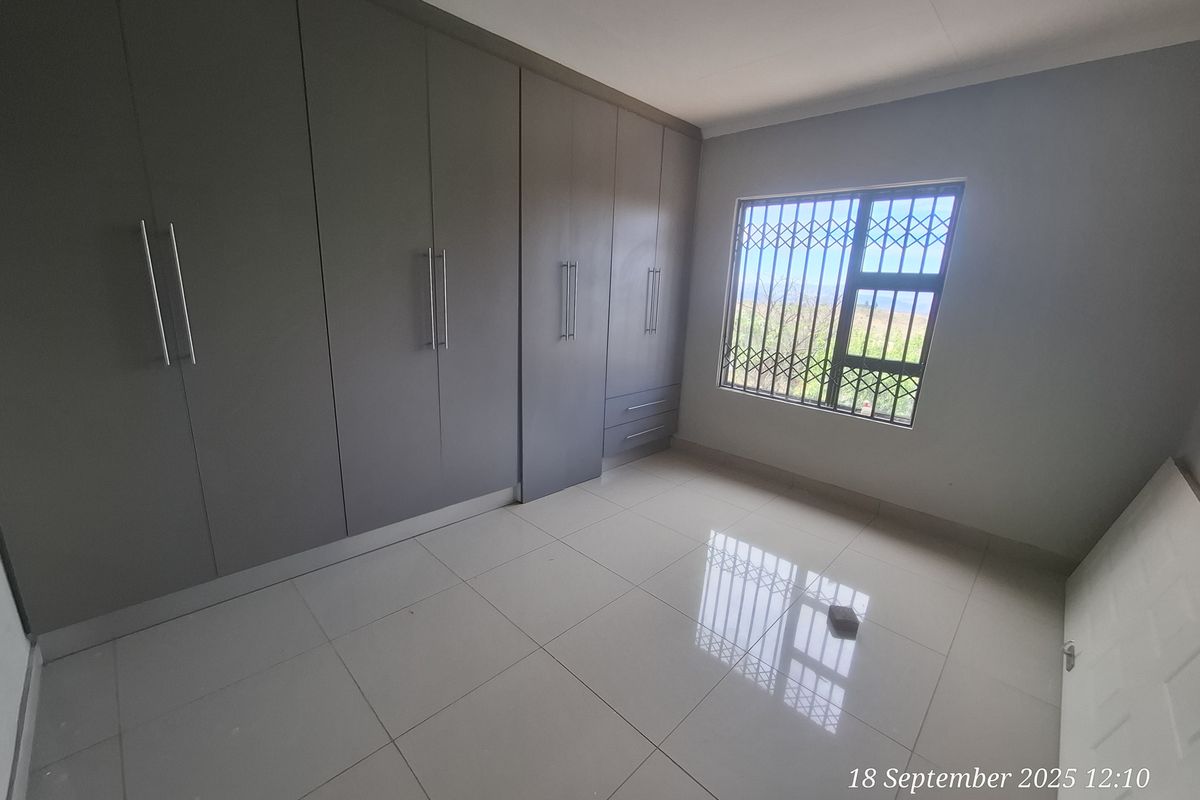 A bright, empty bedroom featuring modern grey built-in cupboards, neutral tiled flooring, and a window fitted with a security gate.