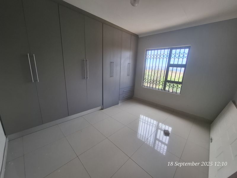 A bright, empty bedroom featuring modern grey built-in cupboards, neutral tiled flooring, and a window fitted with a security gate.