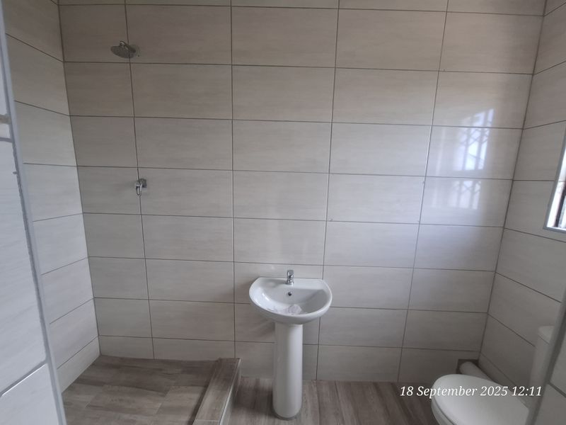 A clean, modern bathroom featuring neutral wall tiles, wood-look flooring, a pedestal basin, and a wall-mounted shower head.