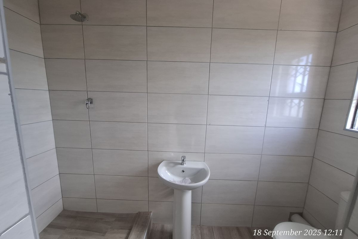 A clean, modern bathroom featuring neutral wall tiles, wood-look flooring, a pedestal basin, and a wall-mounted shower head.