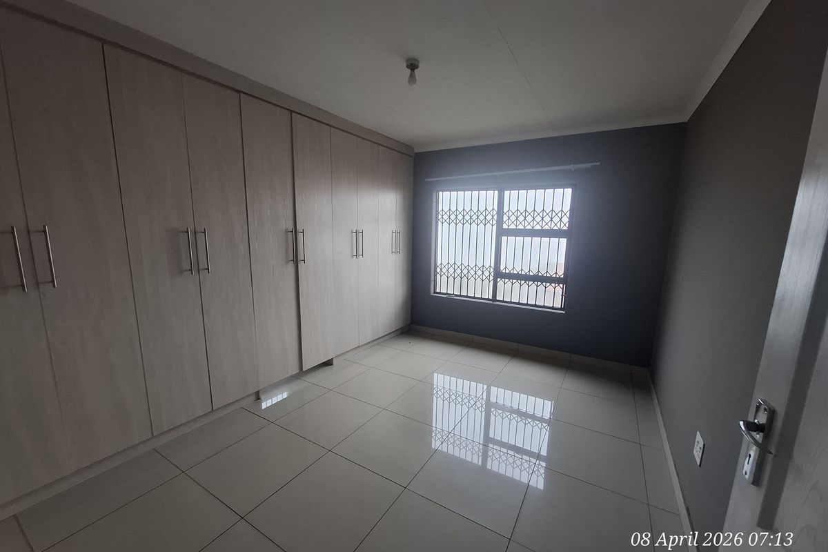 A bright, empty bedroom featuring large built-in wooden wardrobes, tiled flooring, and a window fitted with security bars.