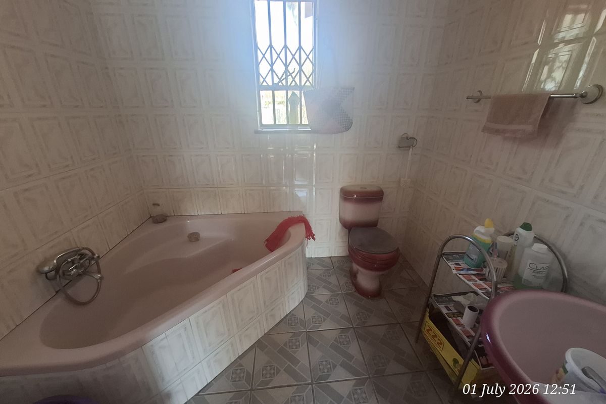 A dated bathroom featuring a corner bathtub, pedestal sink, and toilet, with full-height wall tiling and security bars on the window.