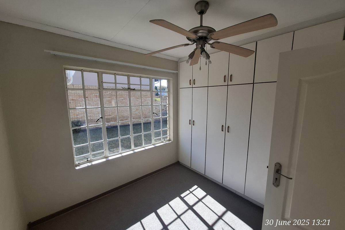 A bright, empty bedroom featuring built-in cupboards, a ceiling fan, and a large steel-framed window overlooking the exterior.