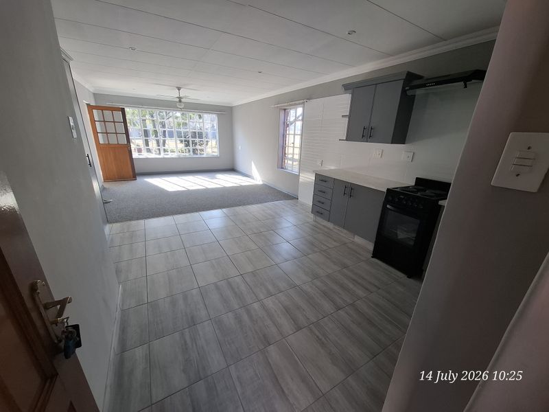 An open-plan living and kitchen area featuring modern grey cabinetry, tiled kitchen flooring, carpeted living space, and natural light through large windows.