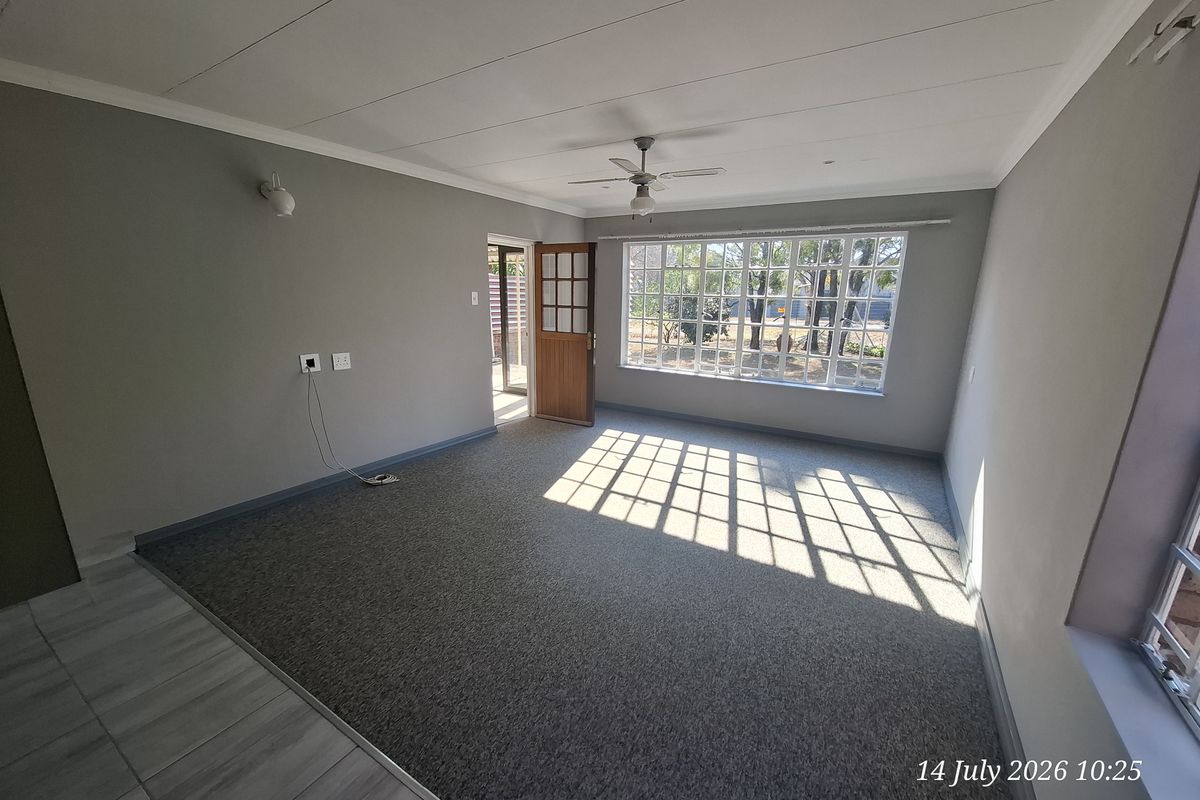A spacious, empty living room featuring carpeted flooring, a ceiling fan, and large grid-pattern windows that provide ample natural light.