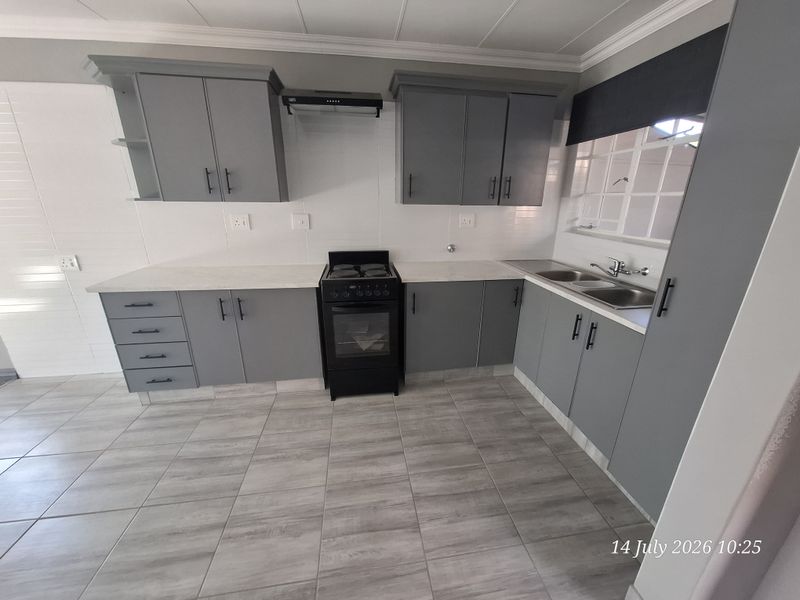 A clean, modern kitchen featuring grey cabinetry, laminate countertops, a freestanding stove with extractor fan, and a double bowl sink with tiled splashback.