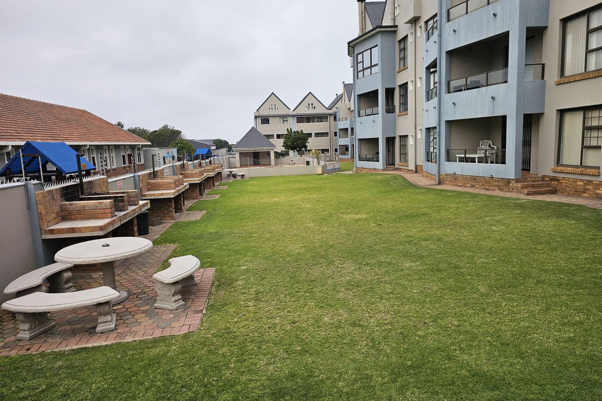 A communal rear garden area featuring multiple built-in brick braai stations, paved seating areas, and well-maintained lawn space adjacent to the apartment building.