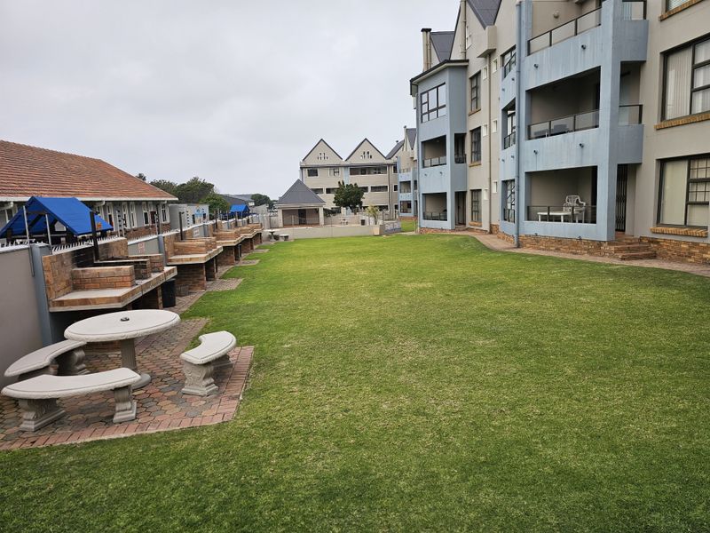 A communal rear garden area featuring multiple built-in brick braai stations, paved seating areas, and well-maintained lawn space adjacent to the apartment building.