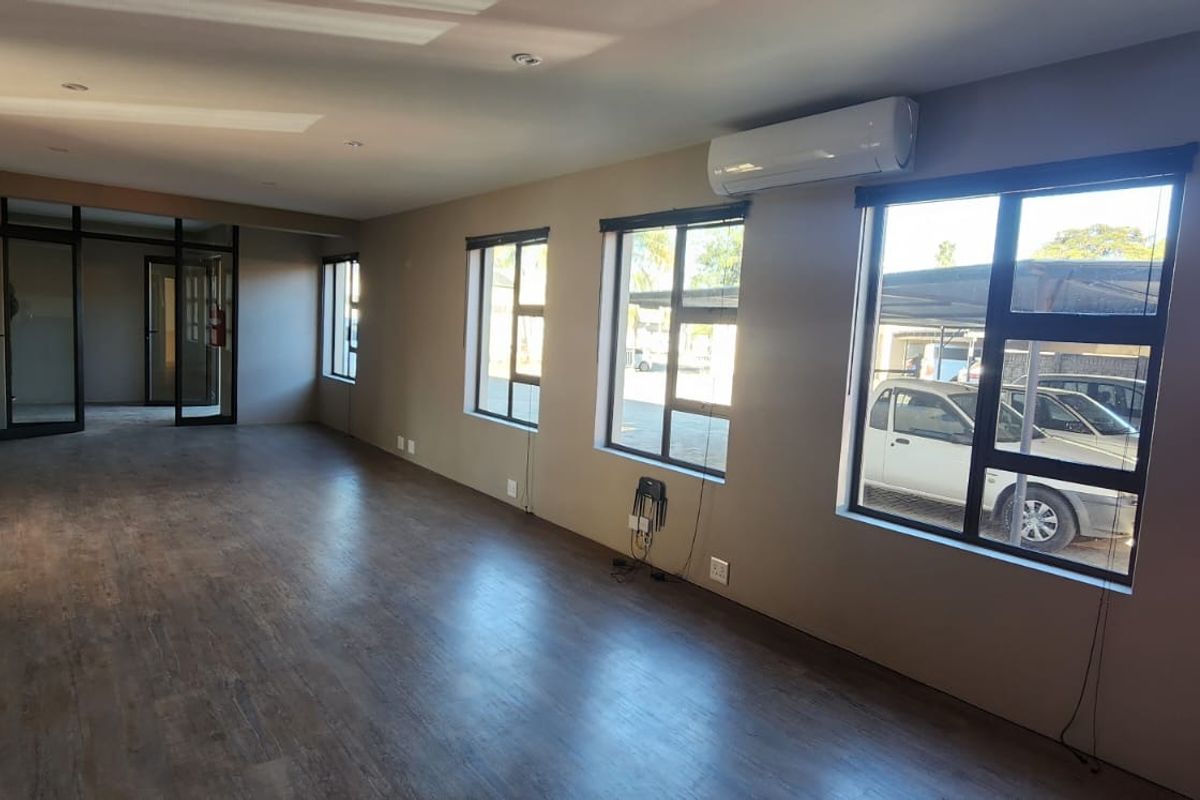 A spacious, open-plan office area featuring laminate flooring, large aluminium windows, and a wall-mounted air conditioning unit.