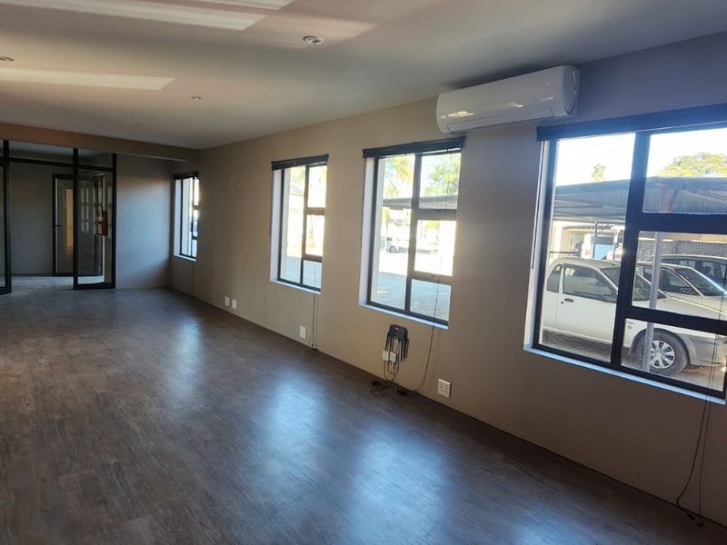 A spacious, open-plan office area featuring laminate flooring, large aluminium windows, and a wall-mounted air conditioning unit.