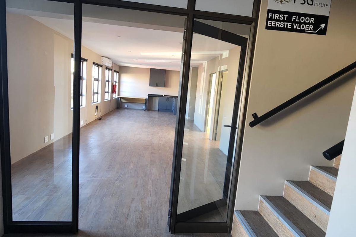 Open-plan office space featuring laminate flooring, a built-in kitchenette, and glass partition access, located near a staircase with a handrail.