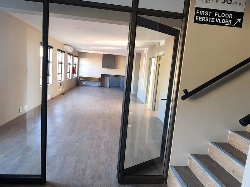 Open-plan office space featuring laminate flooring, a built-in kitchenette, and glass partition access, located near a staircase with a handrail.