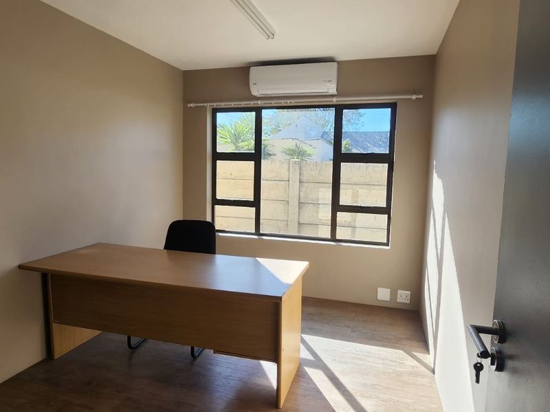 A functional office space featuring laminate flooring, a wall-mounted air conditioning unit, and large windows providing natural light.