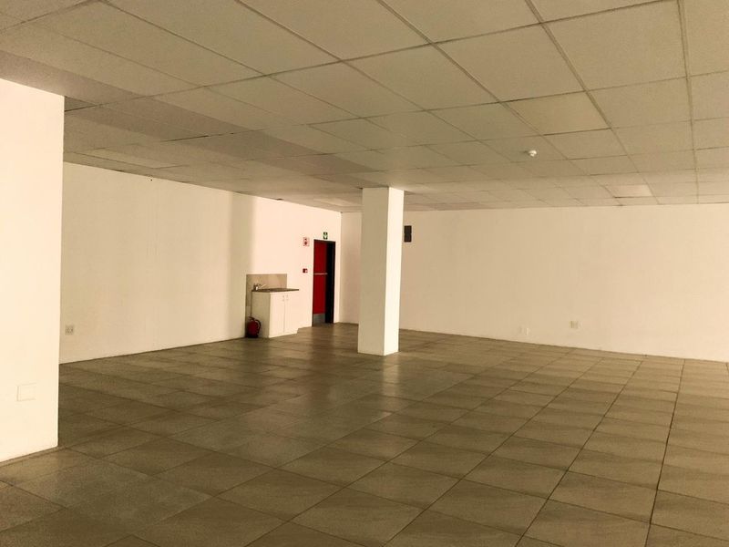 A spacious, empty commercial open-plan office area featuring tiled flooring, a suspended ceiling, and a small kitchenette corner.