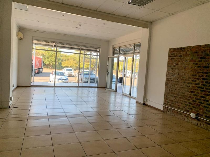 Spacious open-plan commercial unit featuring large glass storefront windows, tiled flooring, an exposed brick feature wall, and a wall-mounted air conditioning unit.