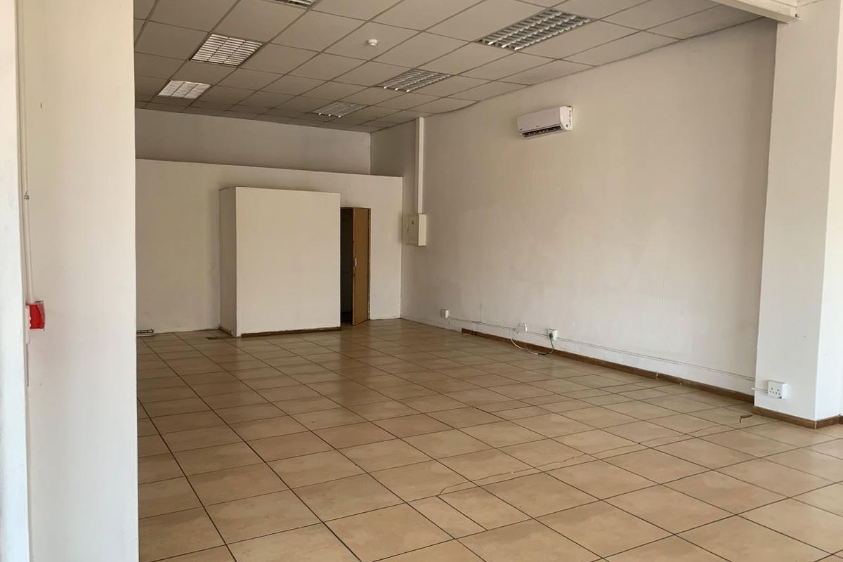An empty, open-plan commercial space featuring ceramic tiled flooring, a suspended ceiling with recessed lighting, and a wall-mounted air conditioning unit.