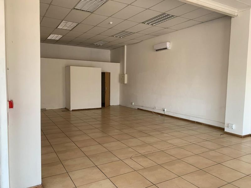 An empty, open-plan commercial space featuring ceramic tiled flooring, a suspended ceiling with recessed lighting, and a wall-mounted air conditioning unit.