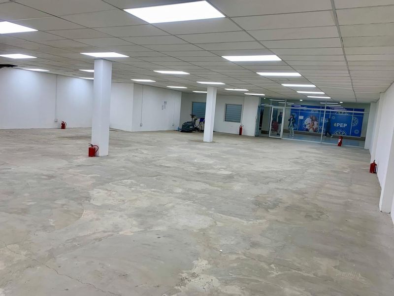 A large, open-plan commercial retail space featuring a suspended ceiling, recessed lighting, structural columns, and a glass storefront entrance.