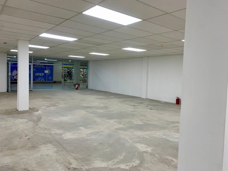 An empty commercial open-plan retail space featuring a suspended ceiling, recessed lighting, and a concrete floor with a glass storefront entrance.