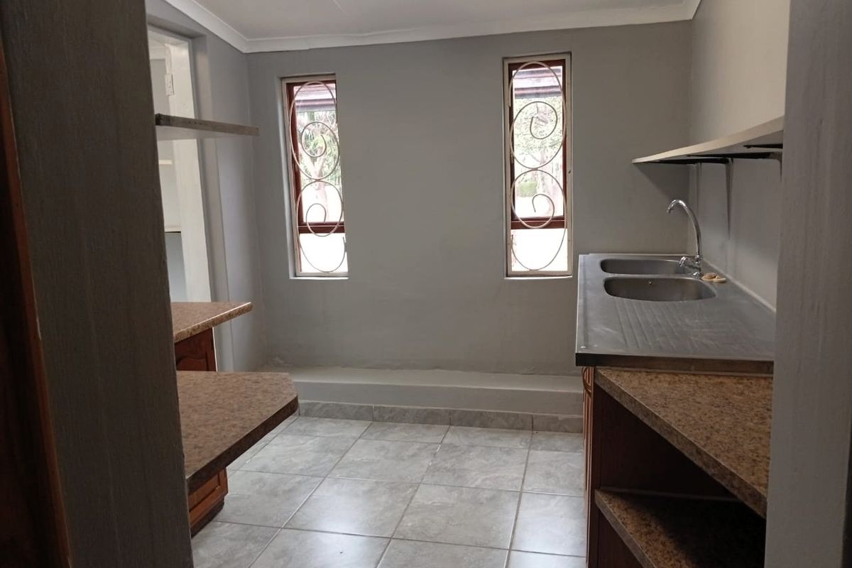 A functional kitchen area featuring tiled flooring, built-in wooden cabinetry, laminate countertops, and a double stainless steel sink with security-barred windows.