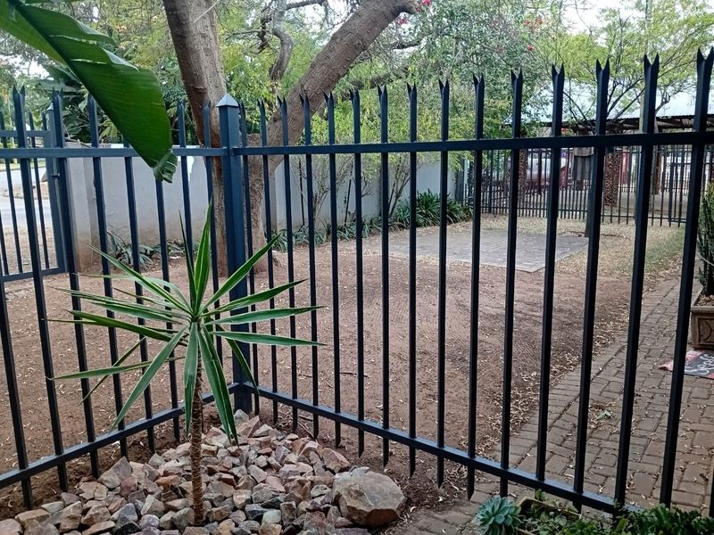 A view of the property exterior featuring secure steel perimeter fencing, a paved walkway, and landscaped garden area.