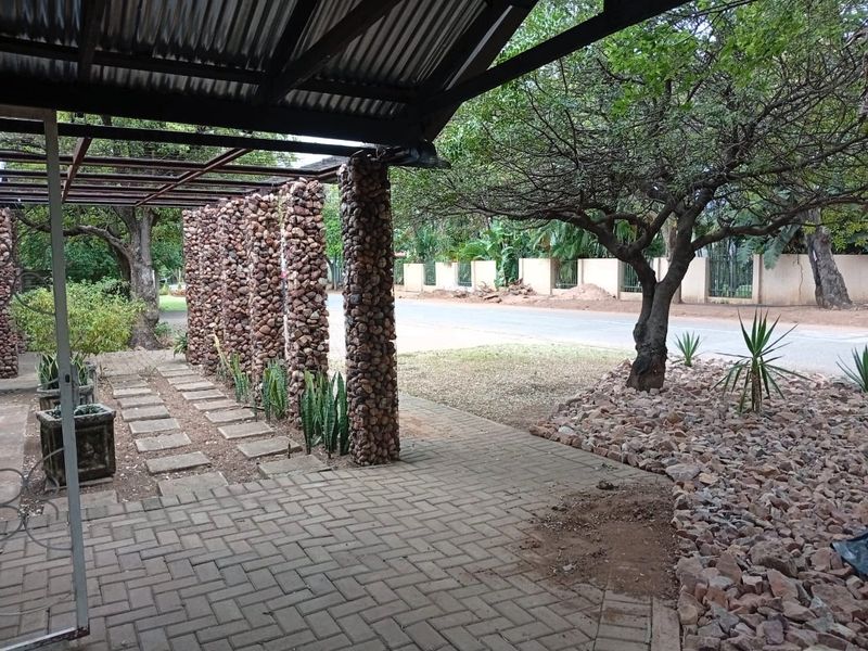 A covered outdoor patio area featuring stone-clad pillars, a corrugated metal roof, and brick paving, overlooking a street-facing garden.