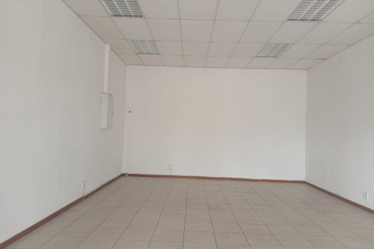 A vacant, open-plan retail space featuring tiled flooring, a suspended ceiling with recessed lighting, and a wall-mounted electrical distribution board.