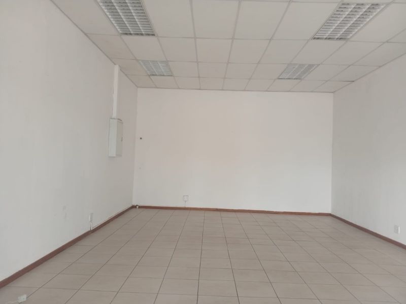 A vacant, open-plan retail space featuring tiled flooring, a suspended ceiling with recessed lighting, and a wall-mounted electrical distribution board.