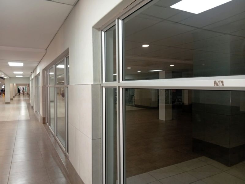 A bright, modern retail hallway featuring tiled flooring, recessed lighting, and large glass storefront panels providing clear visibility into the adjacent vacant retail space.