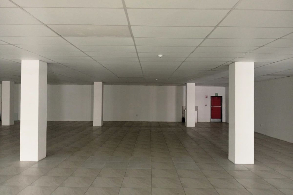 A spacious, empty open-plan retail unit featuring tiled flooring, structural columns, and a suspended ceiling with a rear fire exit.
