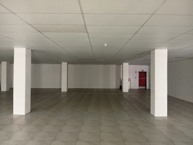A spacious, empty open-plan retail unit featuring tiled flooring, structural columns, and a suspended ceiling with a rear fire exit.
