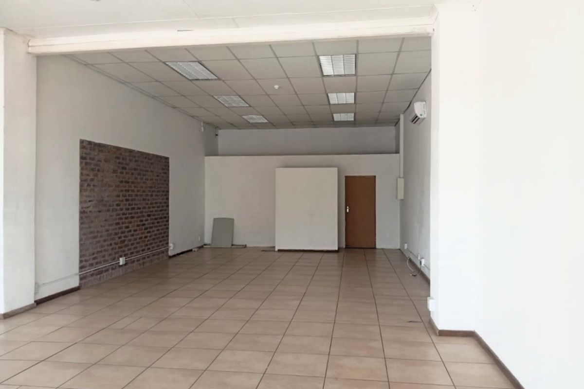 A spacious, empty open-plan retail unit featuring tiled flooring, a suspended ceiling with recessed lighting, and an exposed brick feature wall.